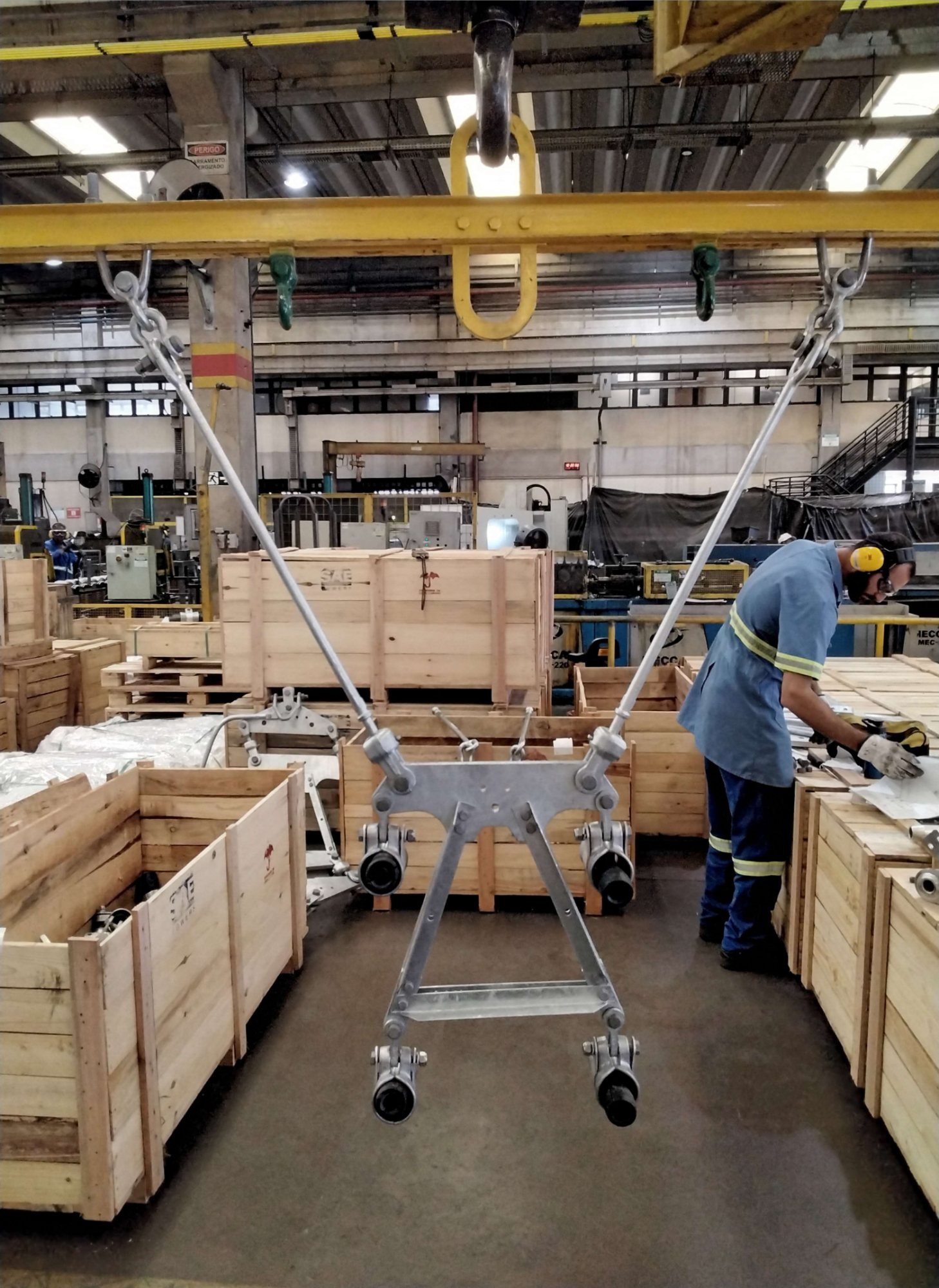 Operário organizando materiais em caixotes de madeira num galpão industrial, com talha suspensa ao fundo